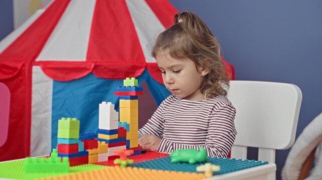 Girl stacking colorful plastic blocks on a textured play table beside a red and white fabric tent in building; concentration.