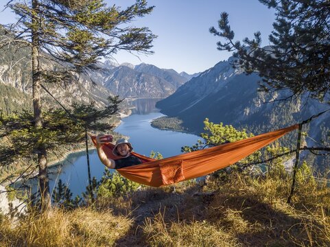 Man taking pictures of himself with mobile phone, hiker sitting in hammock with view of mountains with lake, Plansee, Ammergau Alps, district Reutte, Tyrol, Austria
