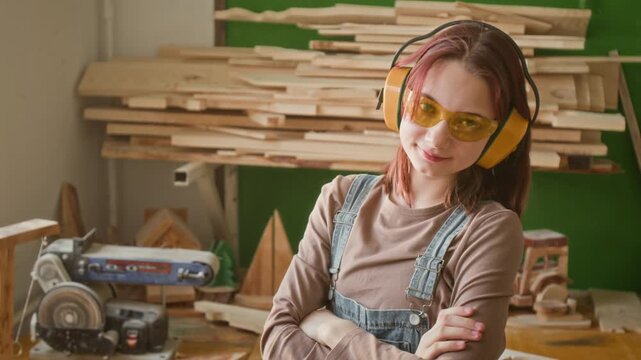 Waist-up portrait of positive red-haired teenage girl wearing yellow safety glasses and earmuffs folding arms and posing for camera in woodworking school