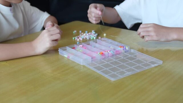 Two children pick colorful beads from a plastic organizer box on a wooden table. Elastic string and sorted beads in pastel and bright colors fill the divided compartments.