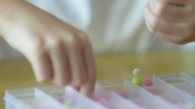 A child threads a small colorful bead onto a thin elastic string with both hands. A plastic bead organizer sits blurred in the foreground below.