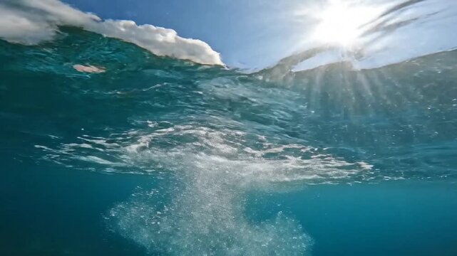 Shimmering underwater perspective looking up at the bright sky with soft white clouds visible through the fluid ripple pattern of the crystal clear ocean surface in high definition.