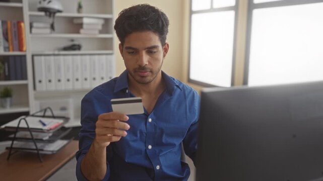 Man holding creditcard while seated at desk, looking at a desktop monitor and keyboard in a modern office building; concentration finance planning.