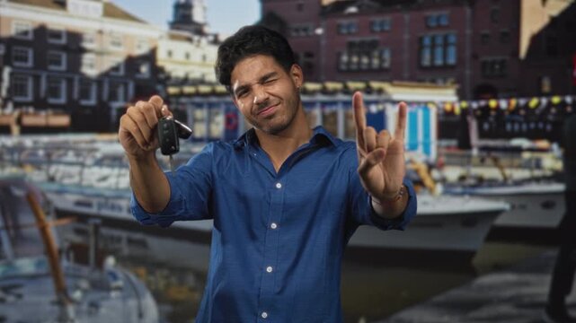 Man holding car keys in right hand, making rock horns gesture on a street by a marina with boats and brick buildings; playful confidence.