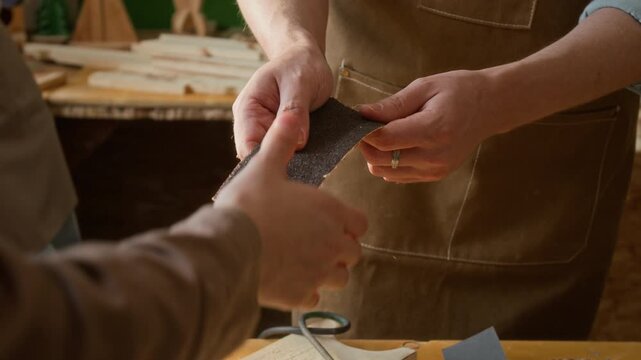 Close-up view of hands of unknown carpenter wearing brown apron showing piece of sandpaper to anonymous children during woodworking class