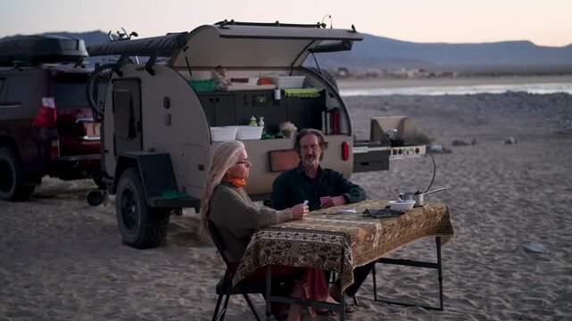 A mature couple plays cards near their teardrop camper by the beach in Baja California as the sun sets on the horizon.
