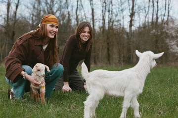 Fototapeta premium Two friends in casual outfits interact with a goat kid on a grassy pasture, capturing a lifestyle outdoors moment with playful animals and companionship in a natural meadow setting, a candid scene of
