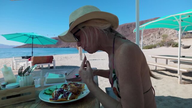 Mature woman sits at a beach stand and enjoys shrimp tacos while overlooking Playa Blanca in Baja California Mexico