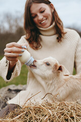 Fototapeta premium Casual outdoor scene of a woman in a sweater bottle feeding a goat kid, capturing a warm rural lifestyle moment and the bond between caregiver and young animal during feeding.