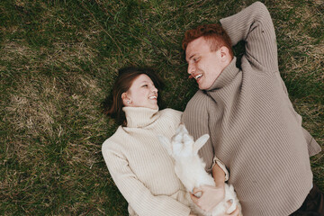 Fototapeta premium Happy couple lying on grass in cozy sweaters, smiling at each other, a small dog between them, outdoor lifestyle moment on a sunny day, intimate candid scene of affection