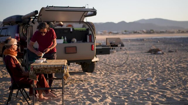 Mature man and woman have dinner outside their camper trailer on the beach at sunset in Baja California, Mexico.