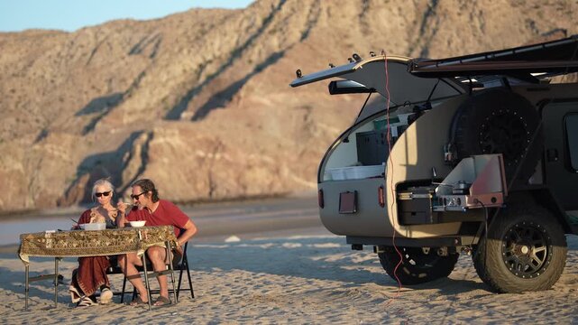 Mature man and woman share a meal by their teardrop camper trailer while camping by the beach in Baja California during sunset.