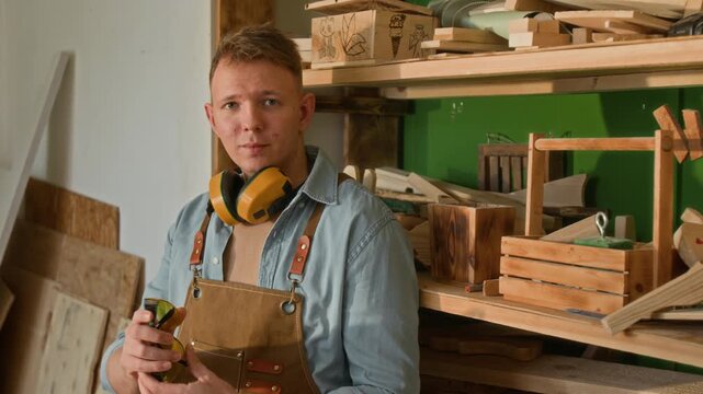 Waist-up portrait of gen Z male carpenter removing protective glasses and posing for camera with crossed arms and smile during workday