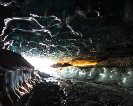 The interior of an ice cave with clear contoured ice on the ceiling, walls of ice and frozen water ripples and waves, a glowing light near the entrance.