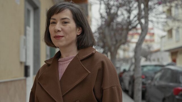 Woman smiling with visible face and brown coat, head slightly turned on a street with parked cars and bare trees; friendly casual warmth.