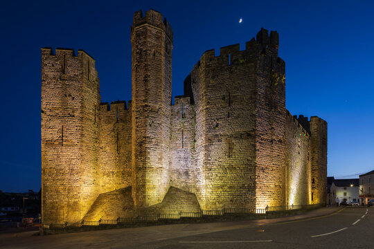 The historic Norman motte and bailey Caernarfon Castle, walls lit up at dusk.