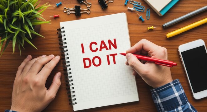Person writing I CAN DO IT in a notebook on a wooden desk.