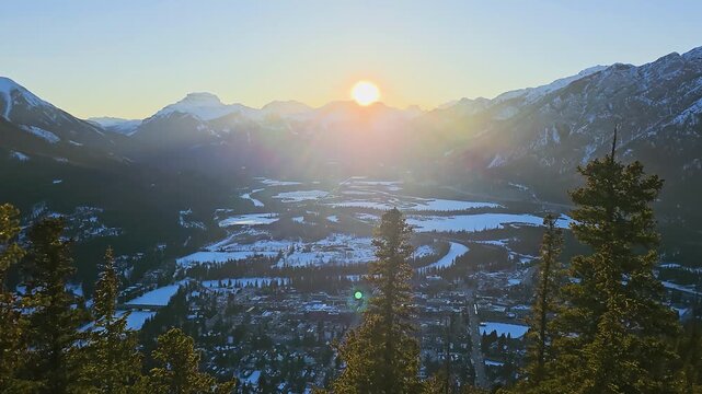 Wide landscape of Banff, Alberta in winter, with the sun setting over the Canadian Rockies, snowy valley floor, pine trees, mountain peaks, scenic town below, and wilderness travel views.