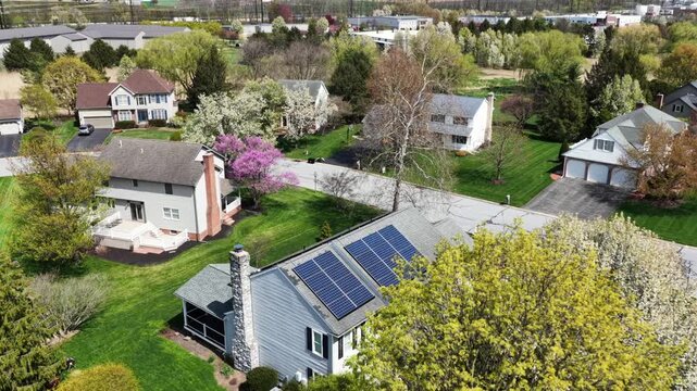Aerial drone view of suburban single-family home with solar panels on roof in United States, surrounded by green lawns, blooming trees and quiet residential streets on sunny spring day.