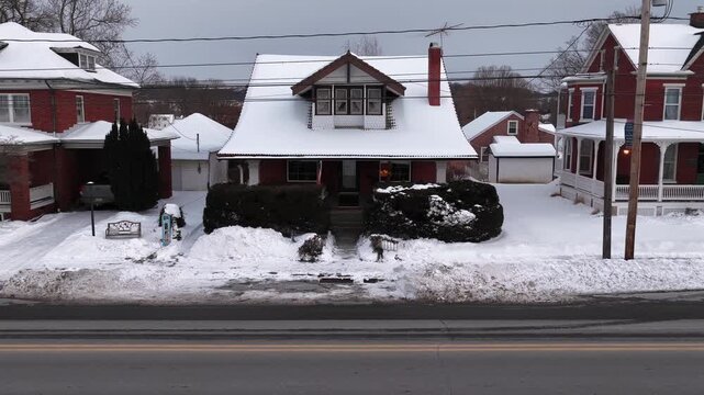 Traditional American house with snow covered roof chimney and front porch in quiet residential neighborhood during winter with parked cars and trees showing small town in United States. Aerial view