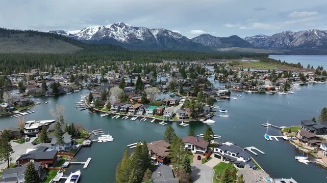 Luxury lakeside neighborhood with canals and mountain backdrop in South Lake Tahoe showing waterfront homes docks and clear alpine water surrounded by forest and snow capped peaks of CA. Aerial wide.