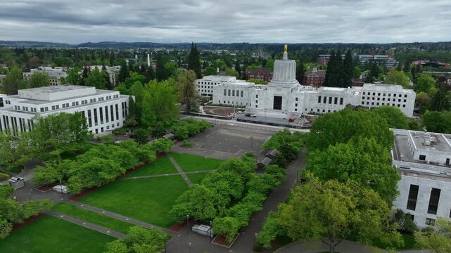 Oregon State Capitol building in Salem surrounded by green park paths and government buildings under cloudy sky. Calm civic atmosphere and administrative center in Pacific Northwest. Aerial approach.