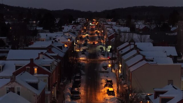 Aerial drone rise over snow-covered small town main street in USA at dusk, with warm streetlights illuminating traffic, row houses and quiet winter atmosphere during holiday season.