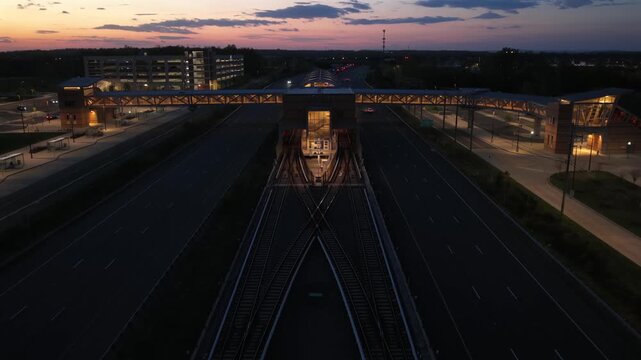 Aerial descent over Ashburn Metro Station in ashburn, Virginia at dusk, showing glowing transit platforms, moving traffic and modern roads beneath colorful sunset sky in suburban USA. Cars on road.