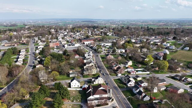 Small town neighborhood in Akron, Pennsylvania with main road, rows of houses and light traffic, surrounded by early spring trees and open land under clear daylight. Aerial wide view.