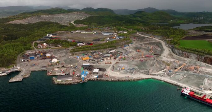 Open-pit quartz diorite stone quarry with heavy machinery, processing plant and cargo ship at port. Aerial drone ascending