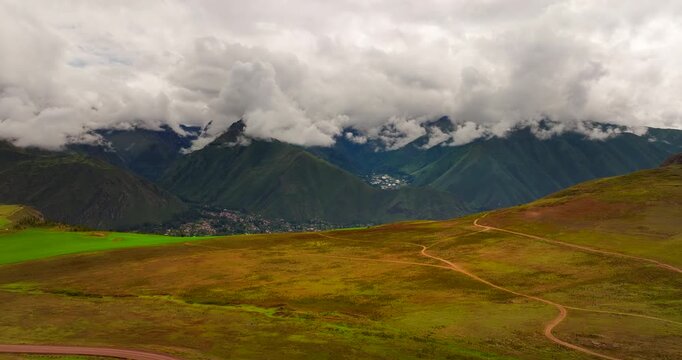 An epic drone shot shows a lone vehicle traveling along a dirt track across the Maras plateau with the Andes mountains of the Sacred Valley and Urubamba Peru, in the background