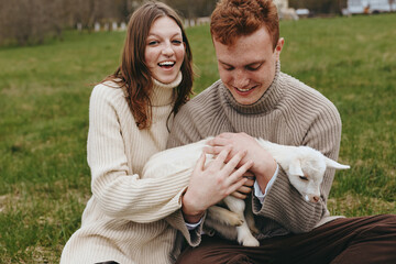 Fototapeta premium Smiling couple outdoors with a goat kid resting in their lap, wearing cozy sweaters. Natural light, warmth, affection, and countryside field vibes convey lifestyle and animal bonding.