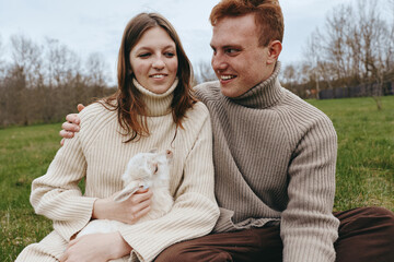 Fototapeta premium Cheerful couple outdoors, warm sweaters, soft sunlight, dog in their arms, relaxed laughter, casual moment, green field backdrop, natural colors, cozy atmosphere, nature
