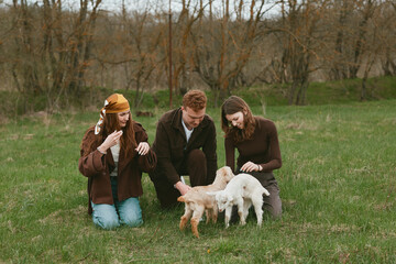 Naklejka premium Three young adults enjoy a spring day outdoors playing with two baby goats on green grass in casual warm clothing creating a relaxed and natural lifestyle moment.
