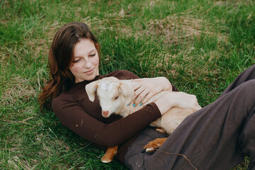 Naklejka premium A goat and kid cuddle with a woman outdoors, a lifestyle moment of bonding and relaxation in a meadow. A tender, calm scene of companionship, nature, and simple joy in bright daylight.