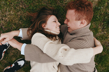 Naklejka premium Smiling couple lying on grass embracing outdoors, cozy sweaters, warm light highlighting their connection. Concept of togetherness, affection, and carefree lifestyle
