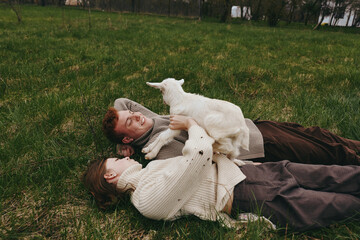 Naklejka premium Lifestyle moment with a couple lying on grass outdoors, a goat kid nearby, they smile and embrace, rustic calm scene, soft lighting, candid everyday joy, harmony between humans and animals.