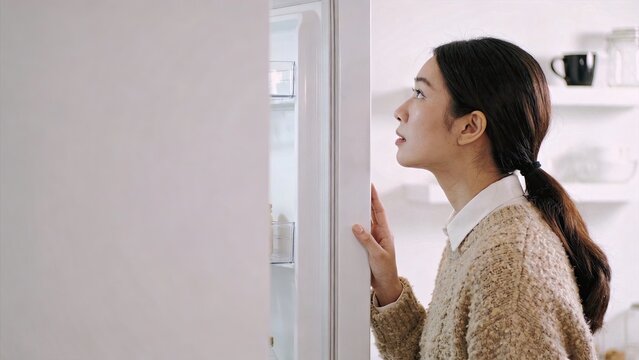 Young Asian woman looking inside open refrigerator at home searching for food or snack kitchen appliance daily life hunger decision making empty fridge lifestyle indoor