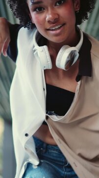 Young smiling dark skinned female with headphones dances outdoors in city environment. Womens joyful expression and casual outfit embody carefree spirit and positive vibes of urban youth culture
