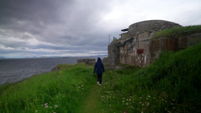 Young woman travels along a grassy path toward the historic ruins of Skrollsvik Fort on the coast of Senja, Norway. Overcast sky creates a moody atmosphere for travel and history themes