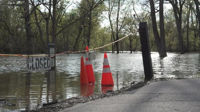 Flooded park entrance and submerged pathway after heavy rainfall. Orange traffic cones and closed signs block access as rising water covers the surrounding area and trees.