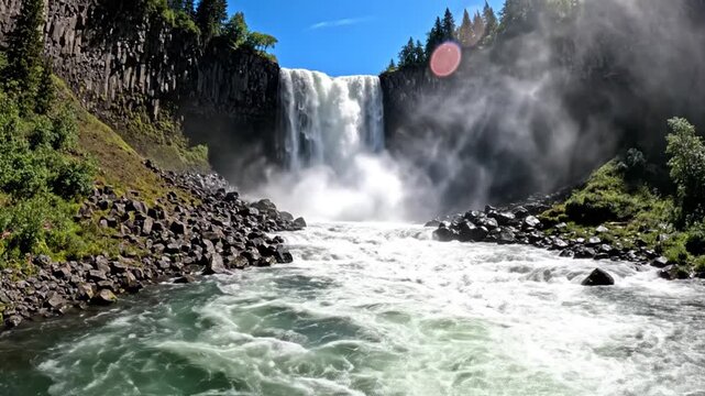 Powerful waterfall plummeting over a basalt rock cliff into a rushing river with mist and spray, surrounded by evergreen forest and jagged rocks under a bright blue sky.