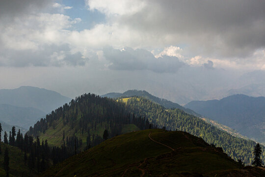 View of Ganga Choti peak with lush green pine forests and a winding trail under a dramatic cloudy sky in Bagh, Azad Kashmir, Pakistan.