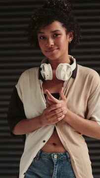 Smiling young woman with headphones around neck, dressed in casual style, standing against black copy space background.cheerful expression exudes confidence and joy,embodying modern and tech lifestyle