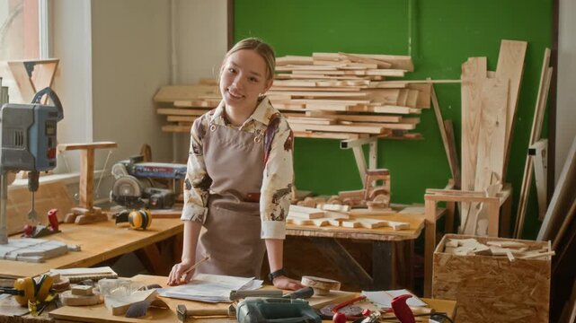 Portrait of happy Caucasian teenage girl smiling for camera drawing sketch while standing at workbench cluttered with carpentry tools in workshop