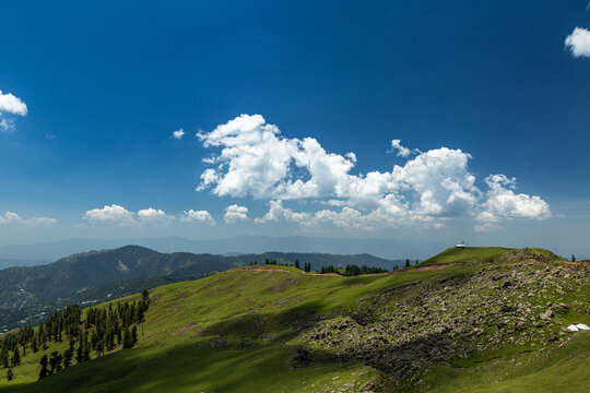 View of Ganga Choti peak featuring lush green slopes and rocky terrain under a bright blue sky with fluffy white clouds in Bagh, Azad Kashmir, Pakistan.