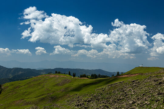 View of the lush green slopes of Ganga Choti peak with rocky terrain and white clouds under a bright blue sky in Bagh, Azad Kashmir, Pakistan.