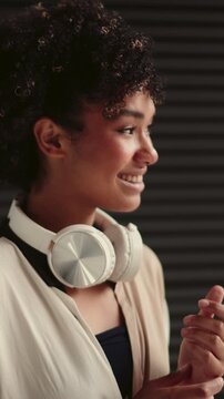 dreamy and shy dark-skinned young woman with headphones around neck, stylishly dressed. stands near black copy space background with relaxed, thoughtful expression, modern and minimalist fashion sense