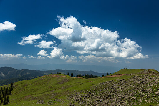 View of the lush green slopes and rocky terrain of Ganga Choti peak under a bright blue sky with white clouds in Bagh, Azad Kashmir, Pakistan.