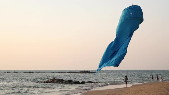 A blue fabric windsock flutters over the sea at sunset over the coastal horizon, slow motion